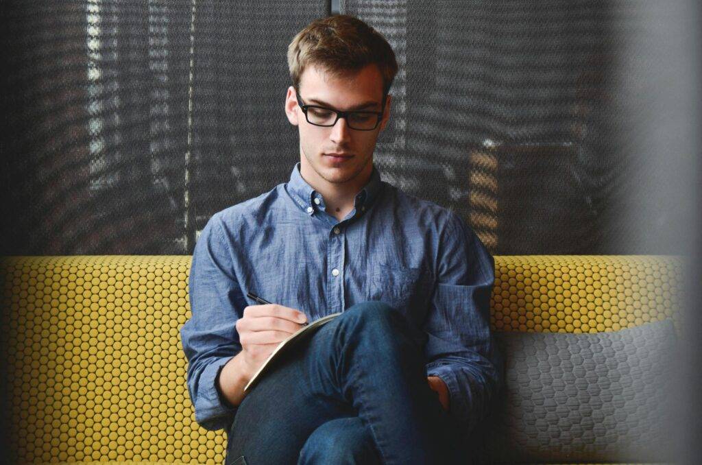 A focused entrepreneur in casual attire sits on a cozy yellow couch, jotting down ideas and strategies for his startup business.