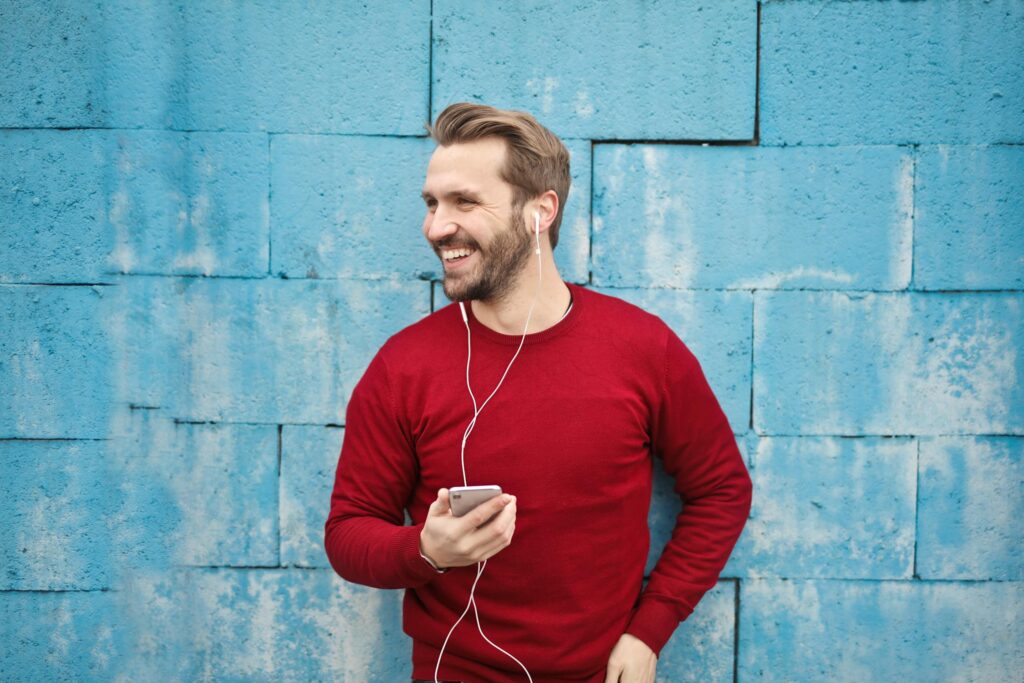 A smiling man in a red sweater stands against a blue wall, holding a smartphone and wearing earbuds, enjoying music.