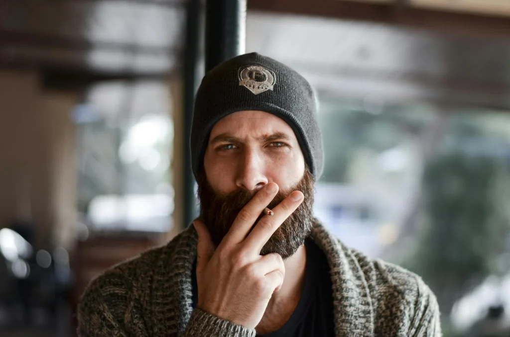 Portrait of a bearded man wearing a beanie and a cozy sweater, thoughtfully smoking a cigarette in a relaxed café setting.