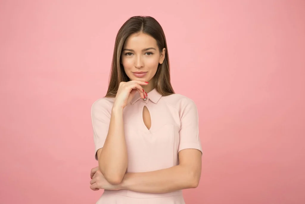 Latina woman with long hair poses confidently in a pale pink outfit against a soft pink background, showcasing a charming and thoughtful expression.