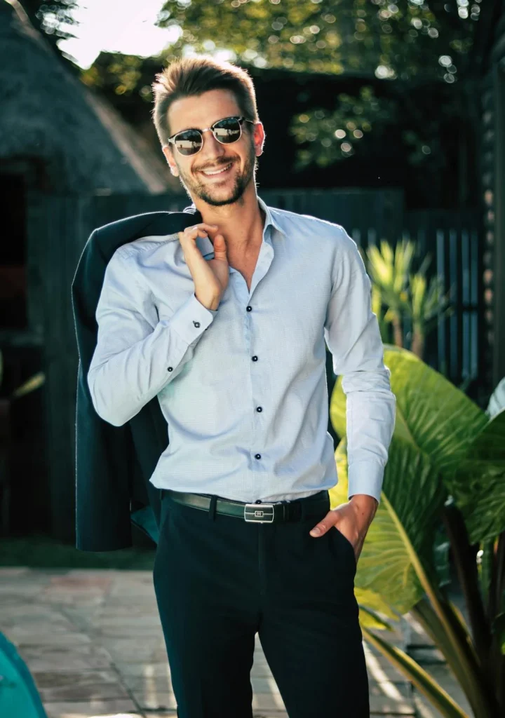 Smiling man in sunglasses wearing a light blue shirt and black pants, exuding confidence against a lush outdoor background.