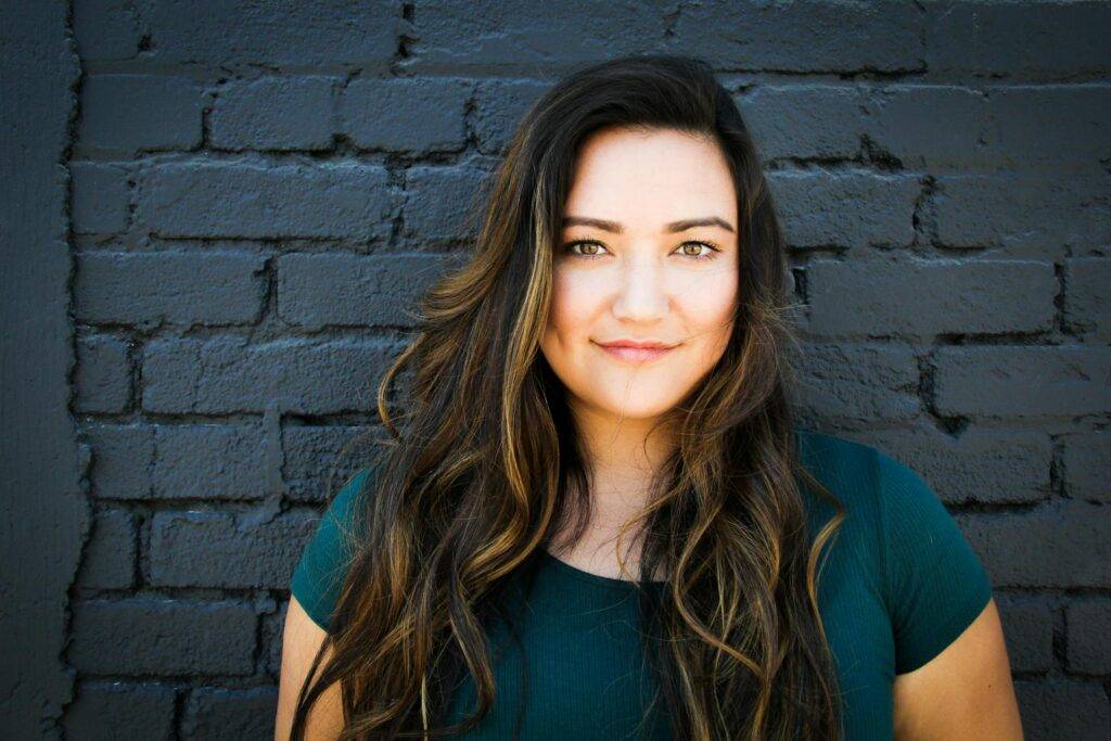 Portrait of a confident young woman with long, wavy hair, smiling against a textured black brick wall, embodying strength and warmth.