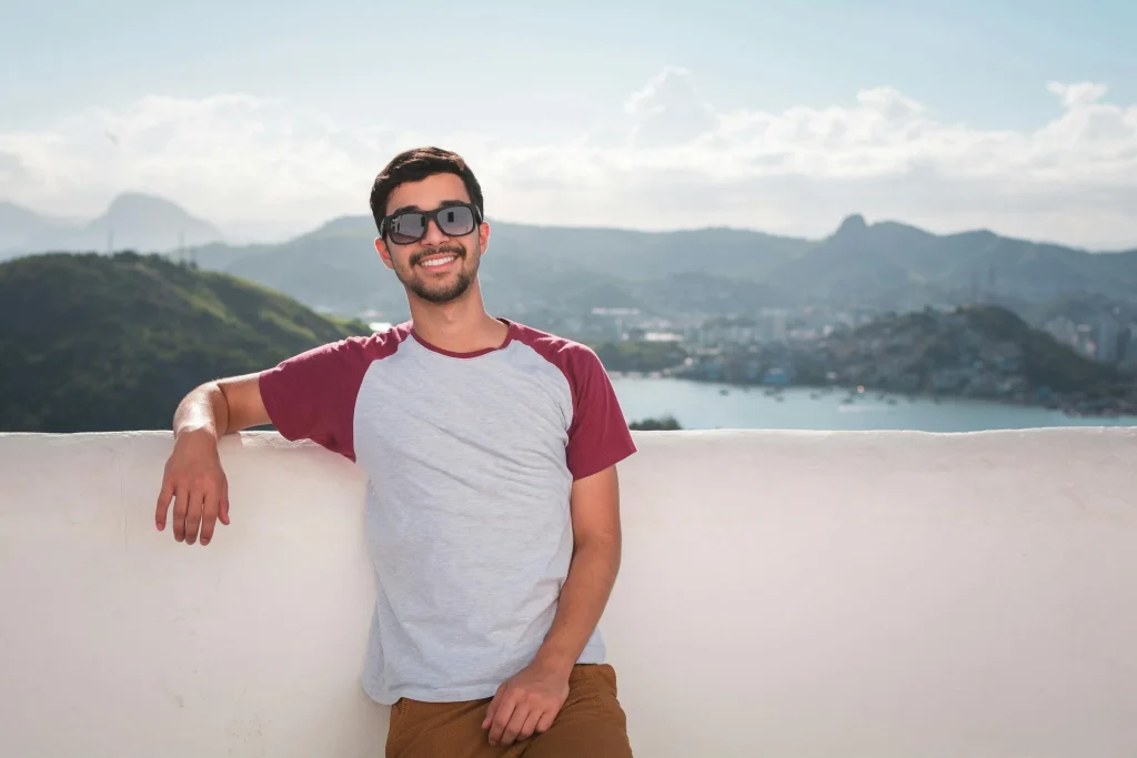 Young man wearing sunglasses and a casual shirt smiles while leaning against a wall, with a scenic view of hills and a bay in the background.