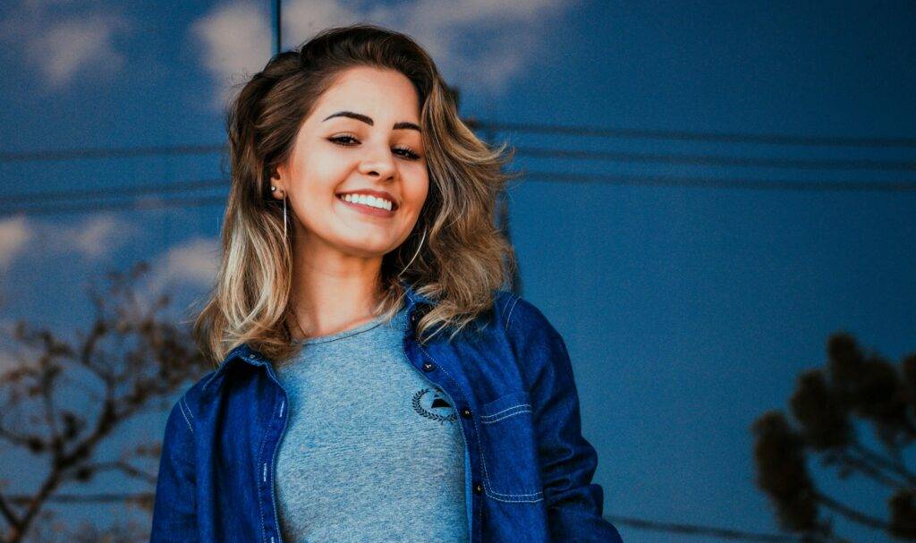 Young woman with long, wavy hair smiles brightly, wearing a denim jacket and a gray shirt, standing against a reflective blue background.