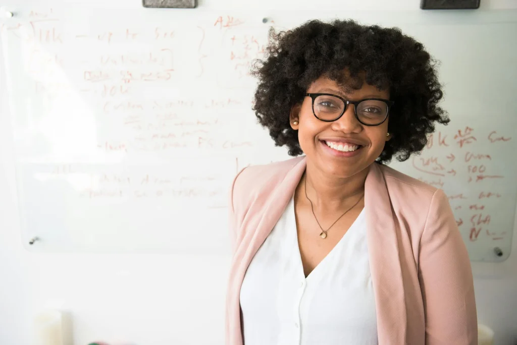 Smiling young woman with curly hair wearing glasses and a blazer stands confidently in front of a whiteboard with notes, showcasing a positive work environment.