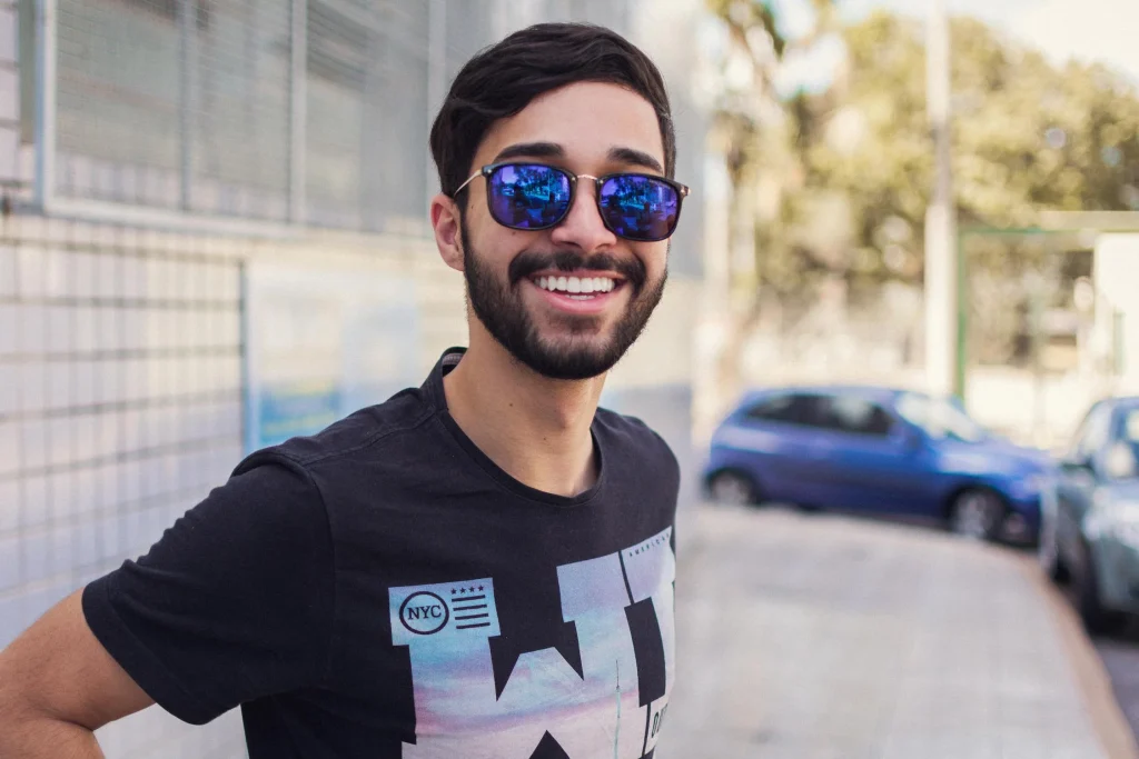 Smiling young man wearing sunglasses and a graphic t-shirt, standing outdoors with cars in the background, radiating a cheerful vibe.