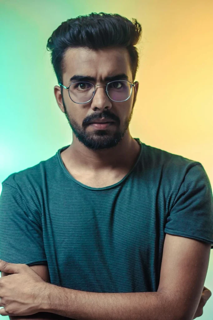 A young man with glasses and a serious expression poses against a colorful background, showcasing a modern and stylish look.