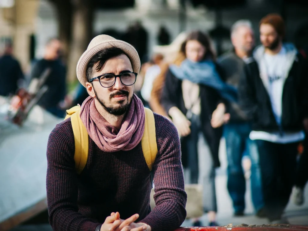 A stylish man in a hat and scarf sits pensively outdoors, surrounded by blurred figures, capturing a moment of urban life and contemplation.