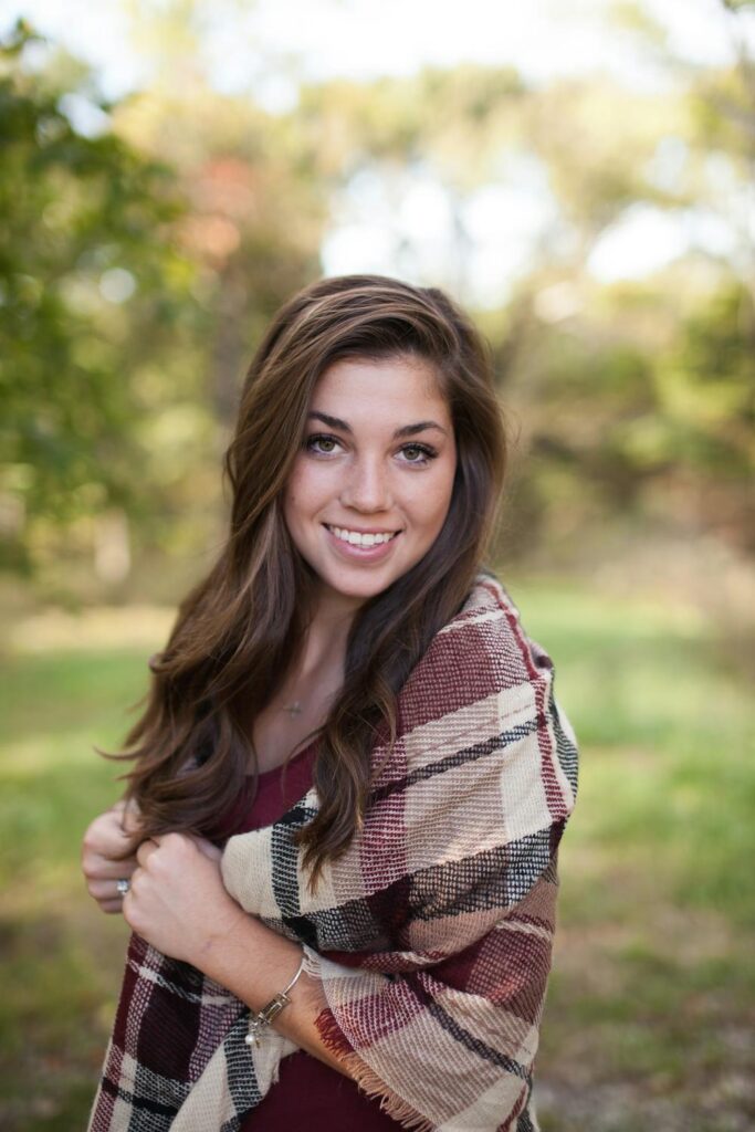 A young woman smiles warmly while wrapped in a plaid shawl, set against a natural, sunlit background of trees and greenery.