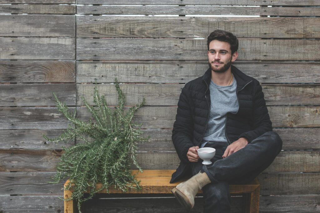 A young man with a beard sits casually on a wooden bench, holding a cup of coffee, beside a leafy plant against a rustic wooden backdrop.