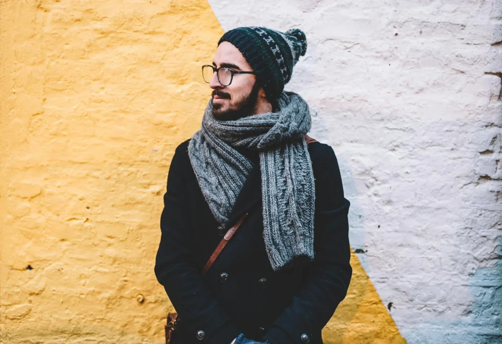 A man with a beard and glasses stands against a yellow and white wall, wearing a warm hat and scarf, exuding a stylish casual vibe.