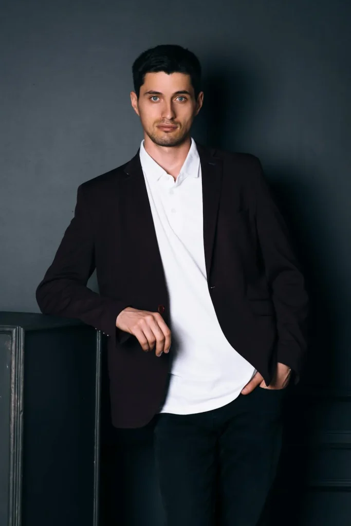 Young man in a stylish blazer and white shirt poses confidently against a dark wall, showcasing a modern fashion aesthetic.