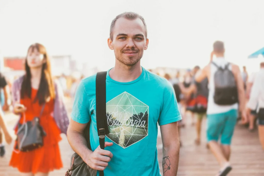 A smiling man in a turquoise shirt stands confidently among a bustling crowd, showcasing a relaxed beach atmosphere.