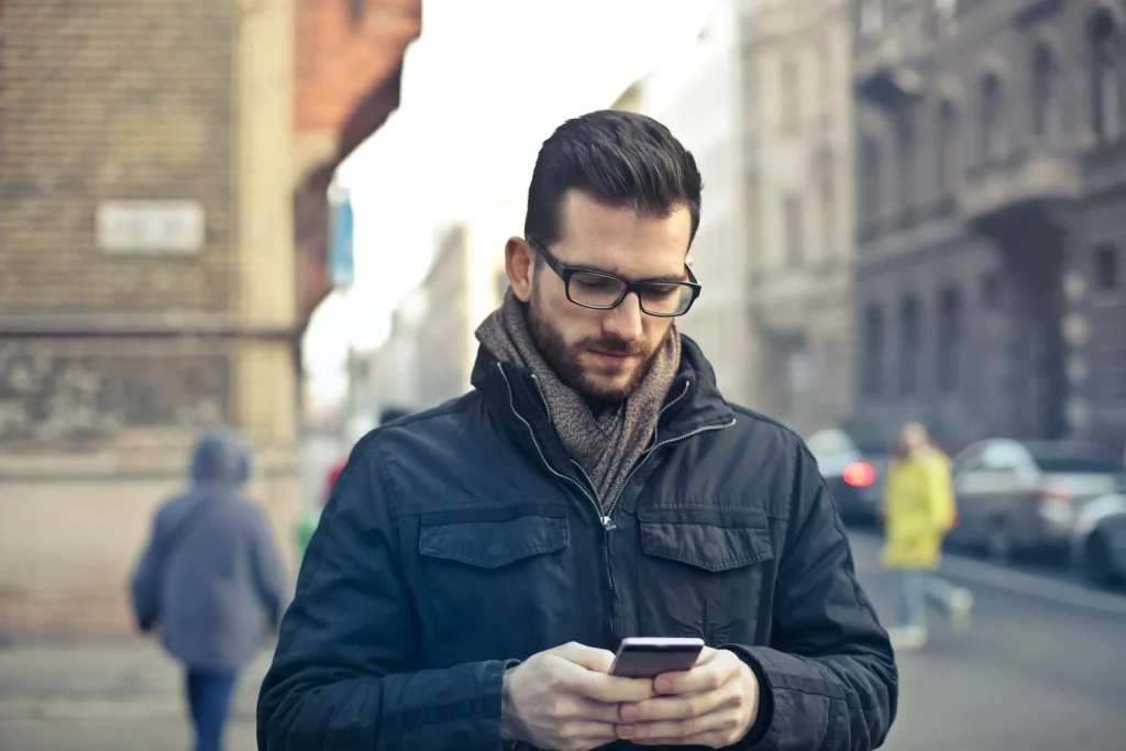 A man in glasses texts on his smartphone while strolling through a city street, surrounded by blurred pedestrians and urban architecture.