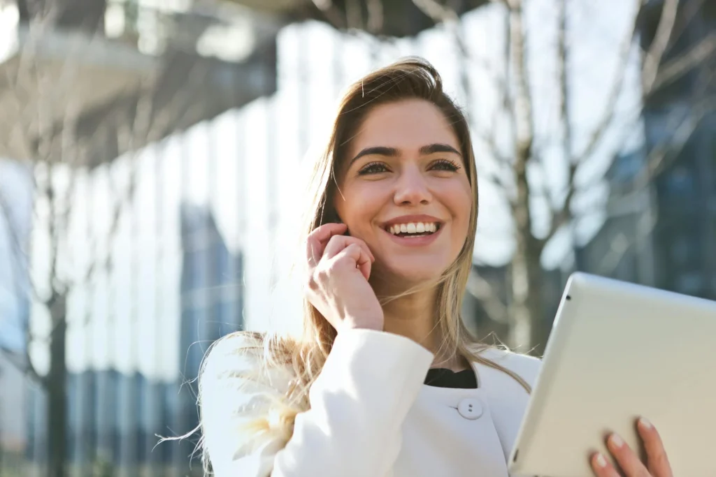 Smiling woman in a white coat holding a tablet while on a phone call, standing outdoors with modern buildings in the background.