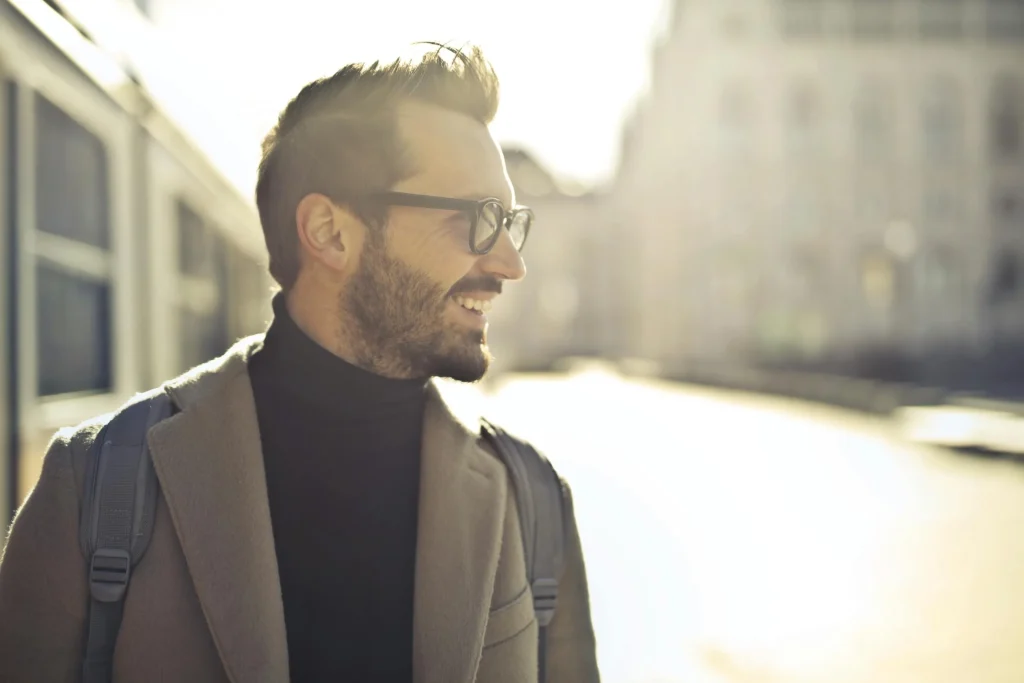 A stylish man with glasses smiles in a sunny urban setting, wearing a brown coat and a backpack, exuding confidence and warmth.