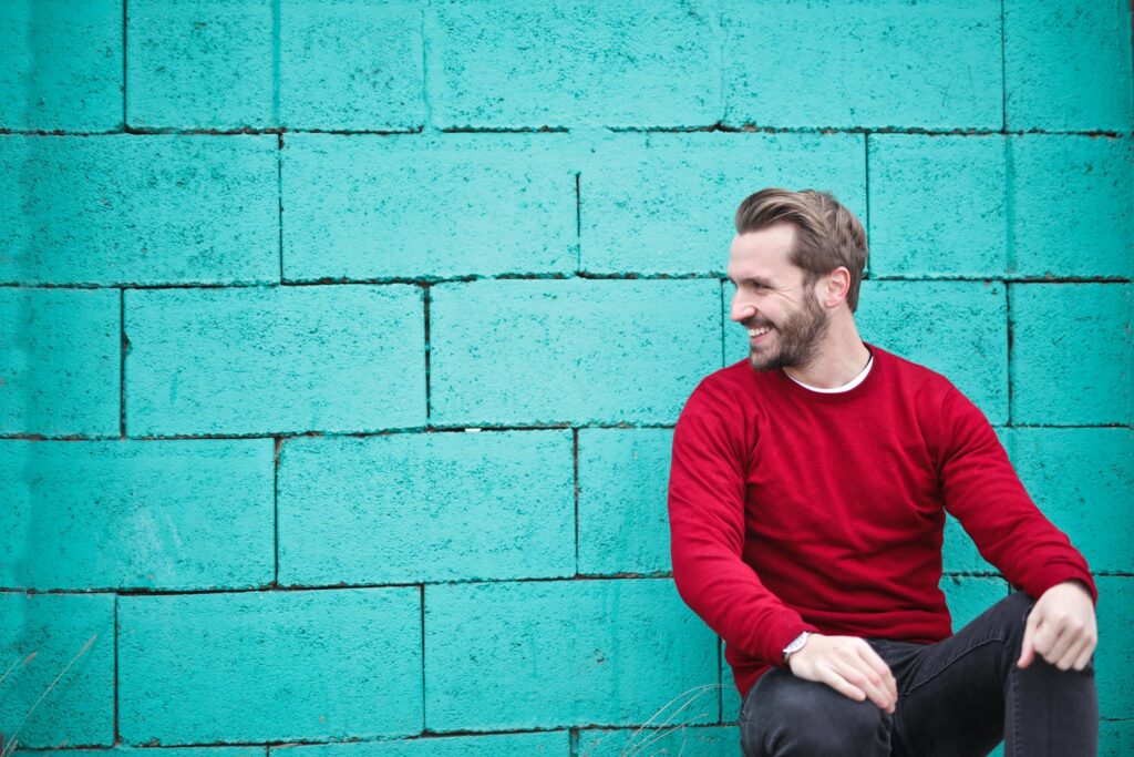 A man in a red sweater smiles while seated against a vibrant teal brick wall, exuding a relaxed and approachable vibe.