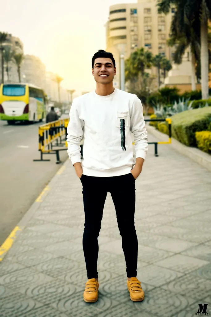 Young man in a white sweatshirt and black pants poses confidently on a city street, with buildings and palm trees in the background.